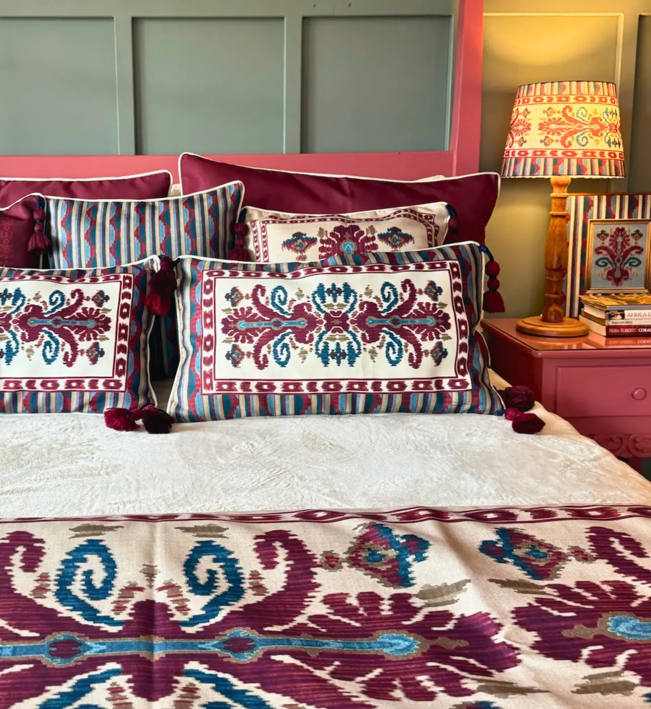 Cama arrumada com almofadas e trilho de cama em padrão ikat nos tons ameixa, azul e creme, ao lado de criado-mudo com luminária acesa.