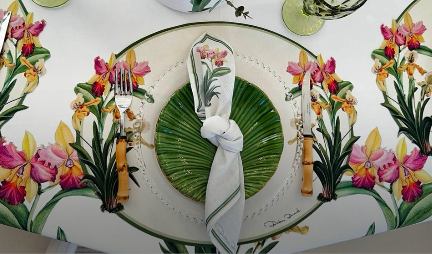 Mesa posta floral com louças, guardanapos e folhas verdes.